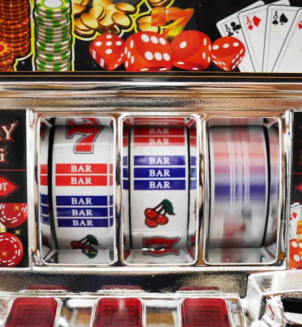 A close-up view of a slot machine with vibrant colors and various gambling symbols such as dice, coins, and playing cards. The reels display a combination of symbols, including bars, cherries, and the number seven. The chrome exterior and colorful details convey the theme of casino and gambling.