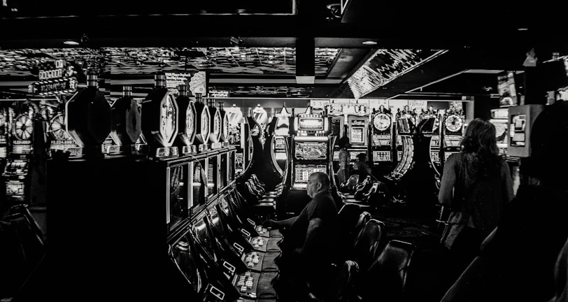 A dimly lit casino filled with rows of slot machines, some occupied by people. The machines' lights contrast with the dark surroundings, creating a vibrant yet subdued atmosphere.