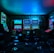 Dimly lit gaming arcade featuring a variety of brightly illuminated slot machines and video gaming screens. Neon signs and colorful displays create a vibrant atmosphere. The arrangement of gaming machines suggests an inviting space for entertainment, with chairs positioned for players.