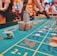 A casino scene with a roulette table features prominently, covered in green felt with colorful numbers and betting squares. Several stacks of poker chips are placed on different numbers, and the hands of several people are visible as they interact with the table. In the background, there are slot machines and more people, creating a vibrant and busy atmosphere.