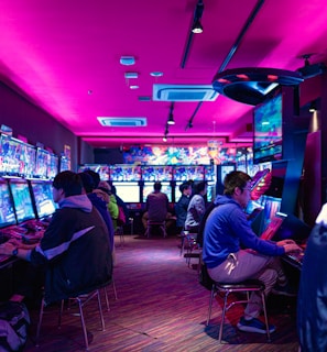 A vibrant arcade with rows of gaming machines adorned with colorful screens. Several people are immersed in playing games, seated on stools in a dimly lit room accented by an intense pink ceiling light. The atmosphere is lively and engaging.