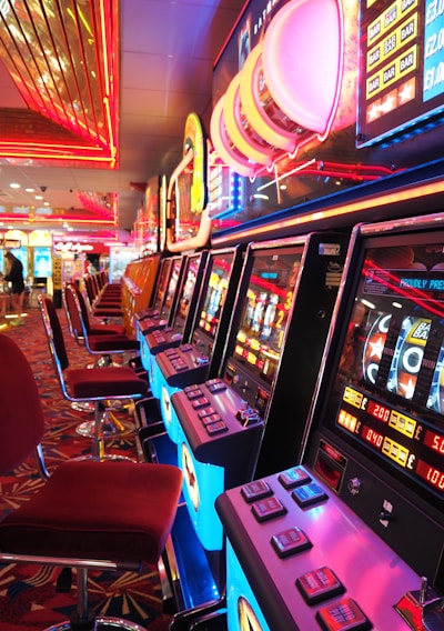 A brightly lit casino interior featuring a row of colorful slot machines. The neon lights create a vibrant atmosphere, with patterned carpeting adding to the decor. Stools are aligned neatly in front of each machine.