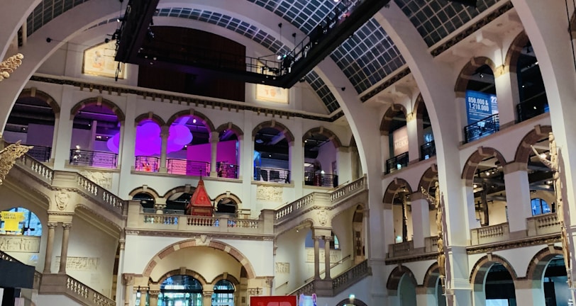 The interior of a large, ornately designed building with arched ceilings and intricate architectural details. The space features gaming machines lined up along the floor, under a high ceiling with a grid pattern. Lighting in purple and white tones enhances the ambiance.