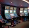 A row of brightly lit slot machines in a casino setting, each with colorful displays and various game themes. The machines are lined up against a dark wall and are unoccupied, with cushioned chairs placed in front of them. The area is dimly lit except for the glow from the screens, creating a vibrant and engaging atmosphere.