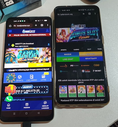 Two smartphones are displayed on a white surface, both showing gambling websites with colorful graphics, text, and various options for logging in and accessing games. The screens are bright and filled with promotional content related to casino and slot games.